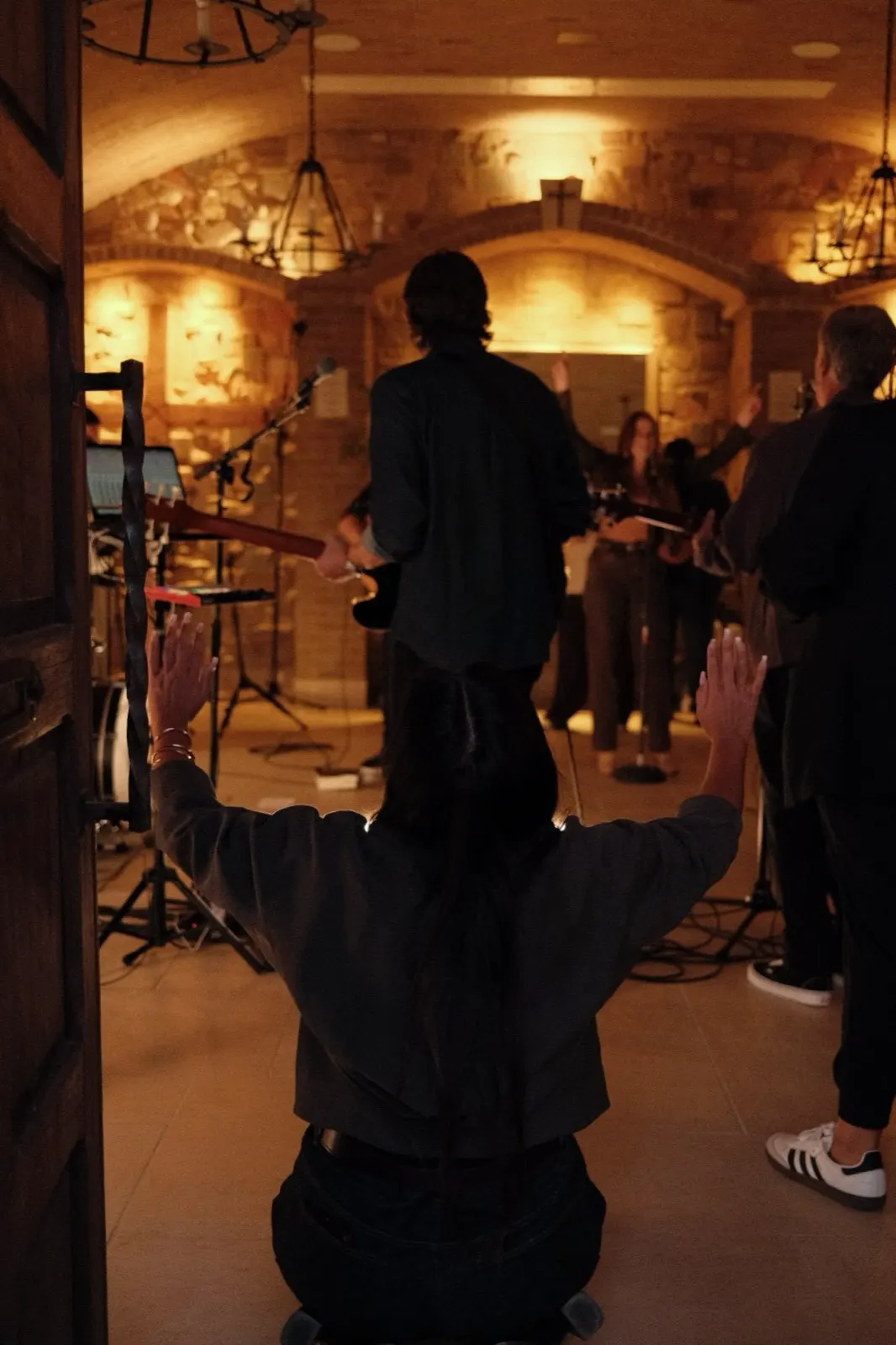 A worshipper kneeling in the doorway with hands raised as the band leads worship beyond, The Hidden Encounter