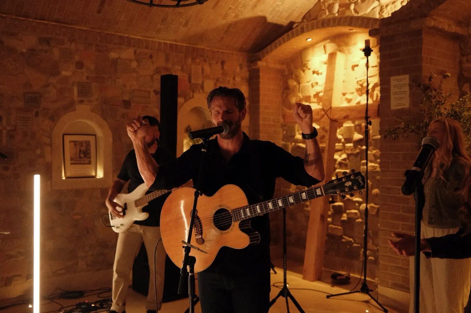 A worship leader singing with hands raised, a wooden cross on the stone wall behind, evening worship at The Hidden Encounter