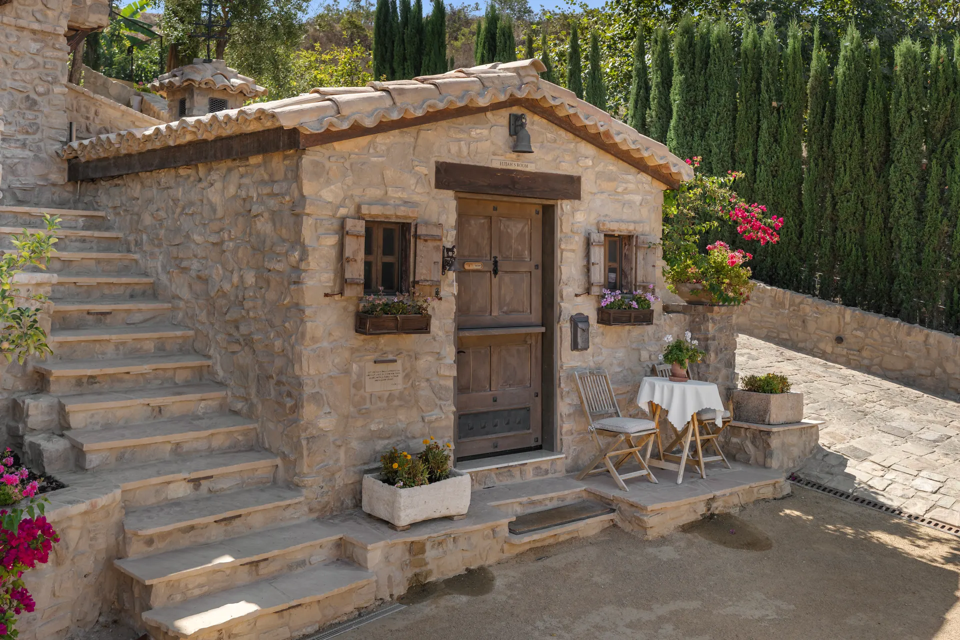 Stone dwelling with an exterior stair climbing past shuttered windows, a small round table and two folding chairs set beside the door