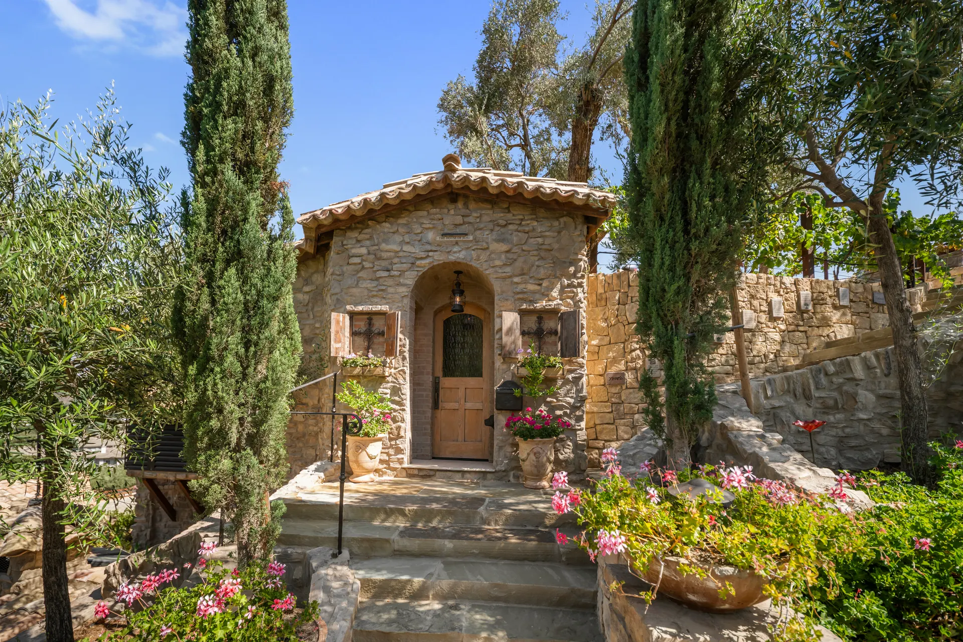 Stone dwelling with an arched wooden door, cypress on either side, geraniums in terracotta pots