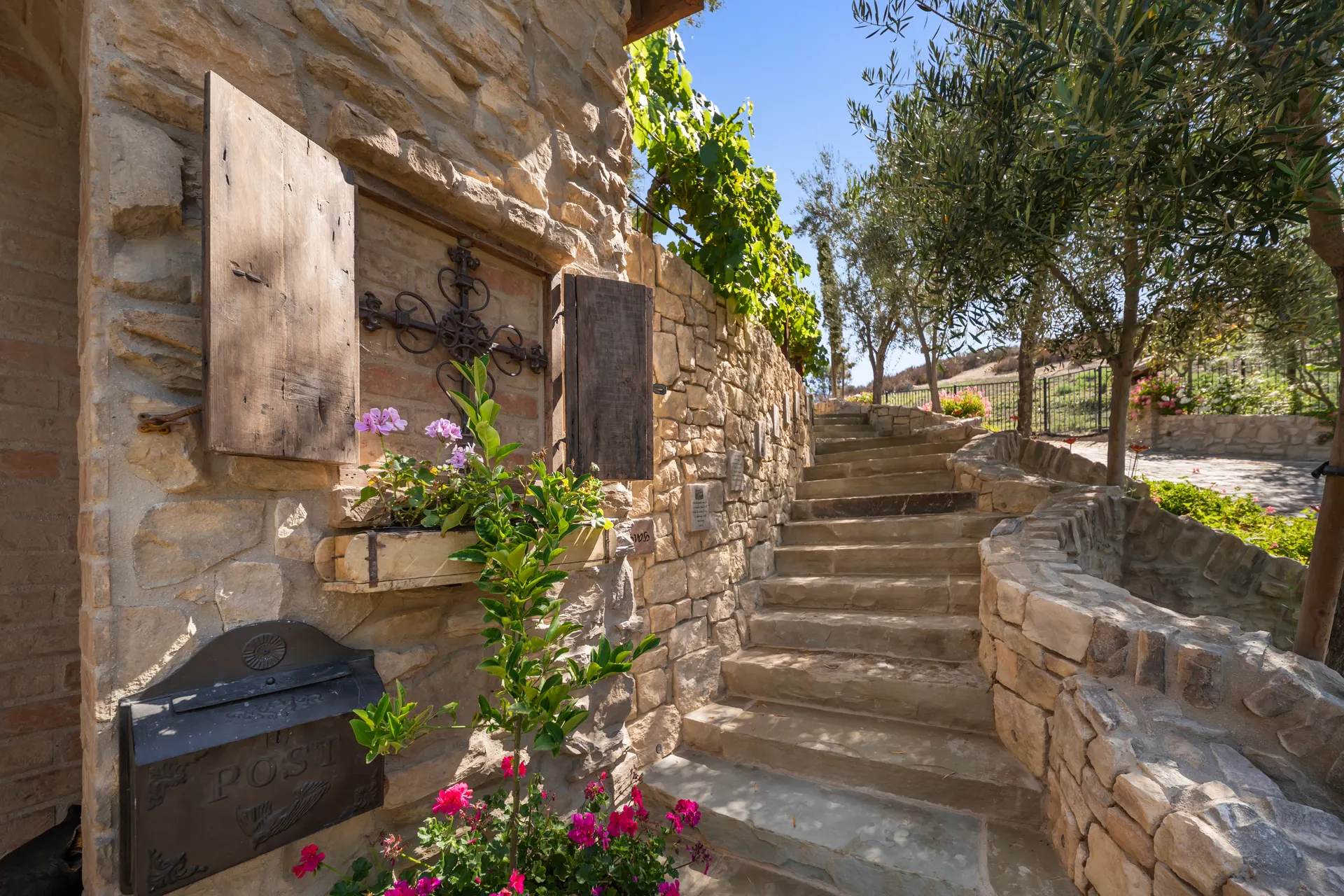 Iron cross mounted on a stone wall beside a cast letterbox and geraniums, stone steps climbing between olive trees