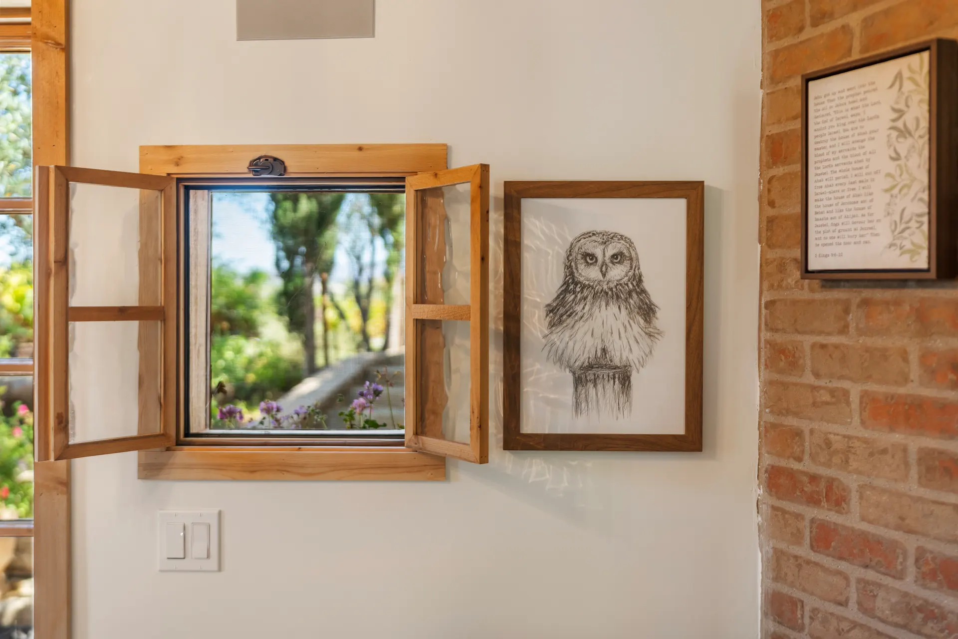 Shuttered window opened to olive trees, framed drawing of an owl on the brick beside it