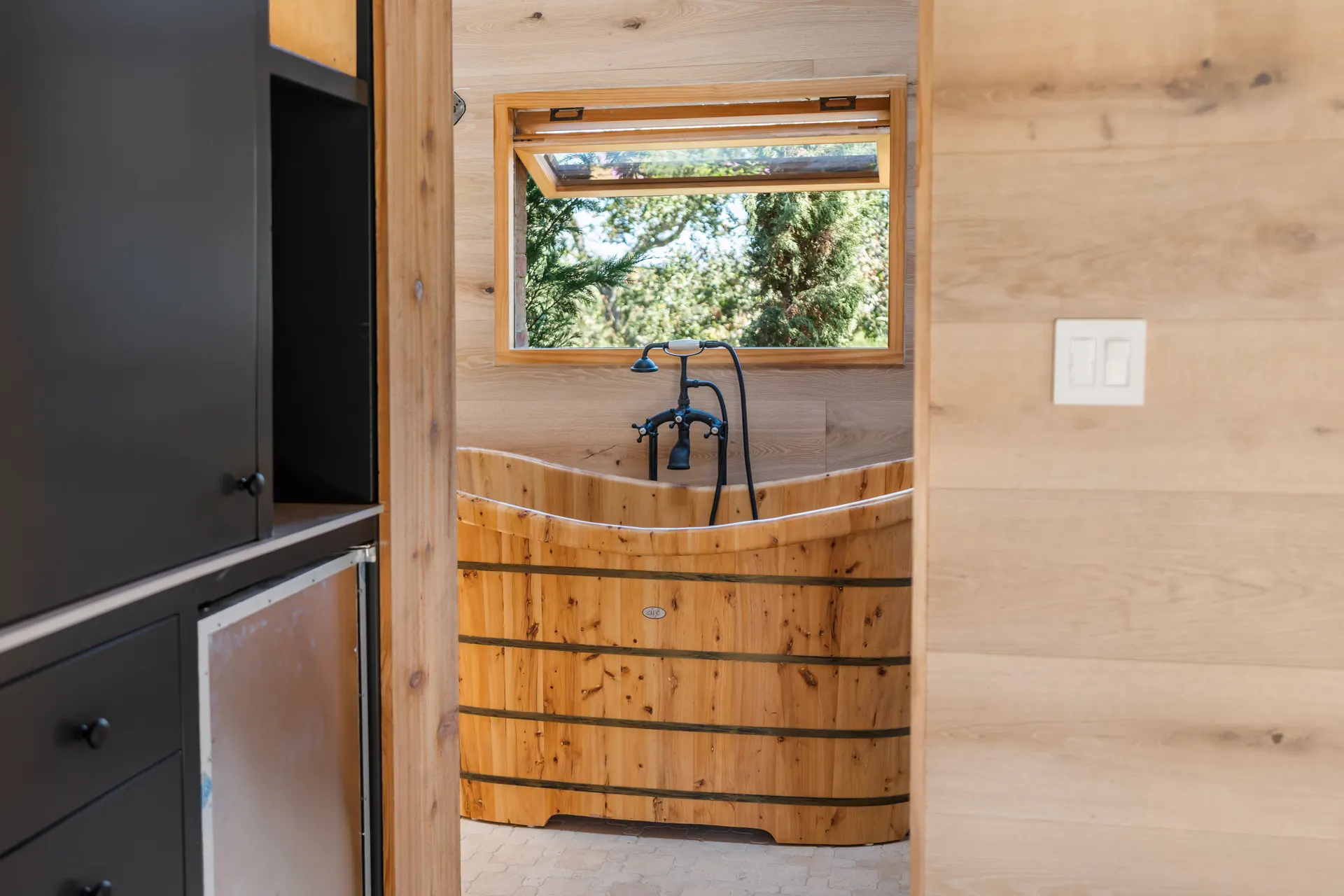 Wooden soaking tub set below a window, plank wall and dark cabinetry alongside