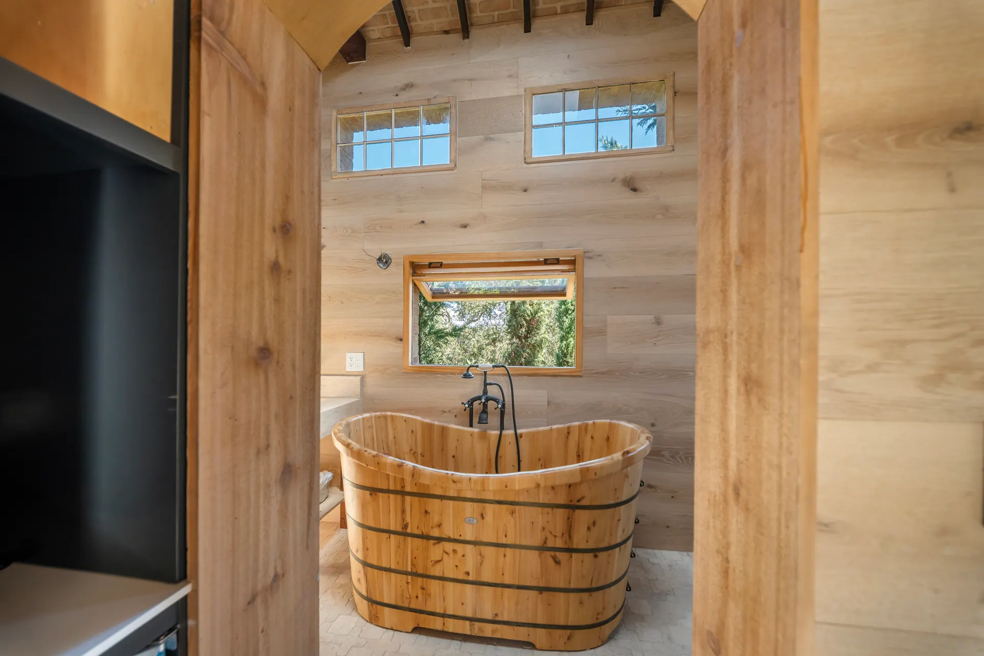 Wooden soaking tub with iron taps beneath a window looking into the trees